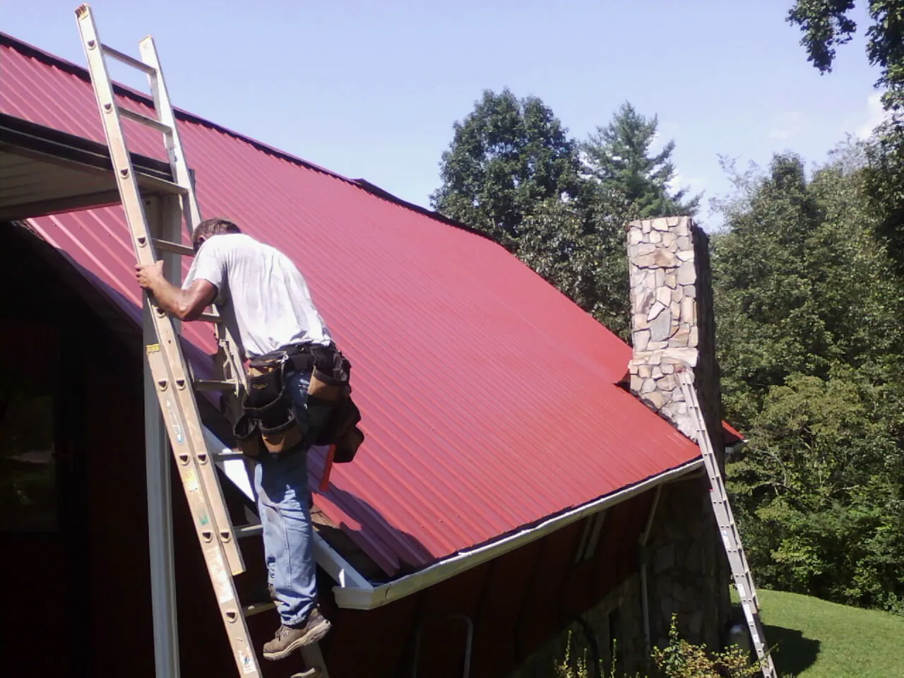 Roofer on a ladder beside a red metal roof and stone chimney — Soffit & Fascia Repair by Roofing Brothers in Bulger